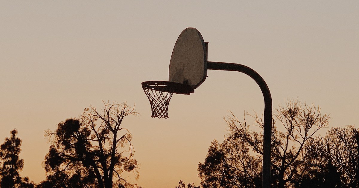 A silhouetted basketball hoop against a dusk sky with trees in the background