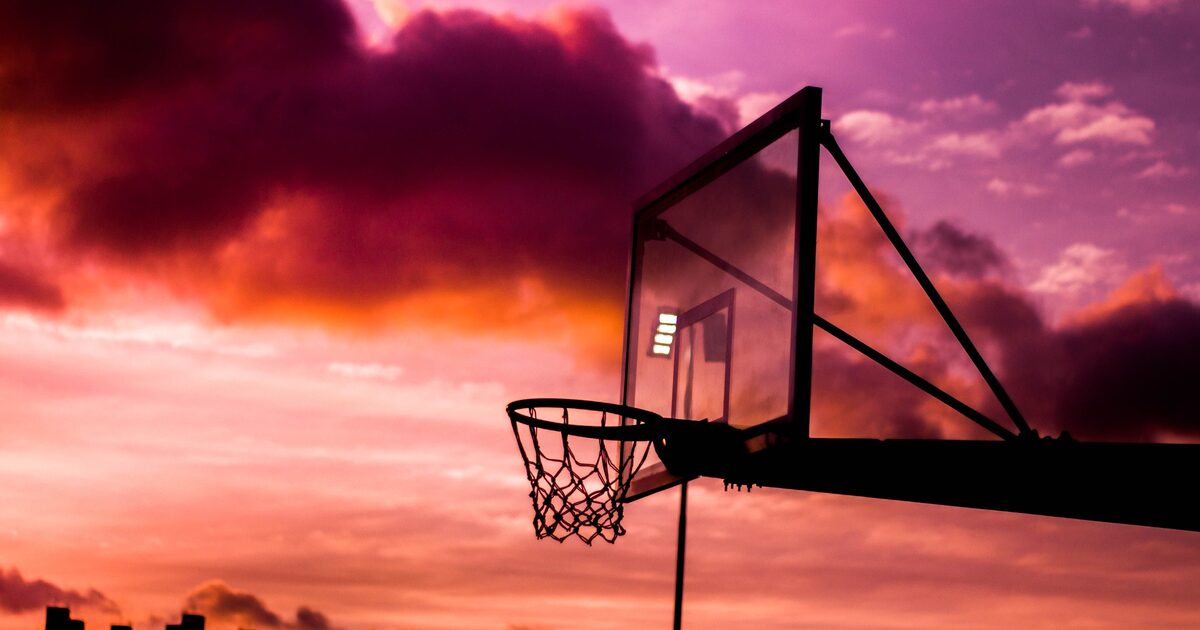 Silhouette of an outdoor basketball hoop against a vivid orange and purple sunset sky