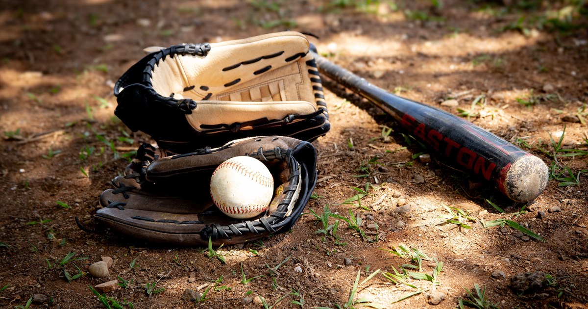 A worn leather baseball glove, a baseball, and a wooden bat on a dirt infield in warm light